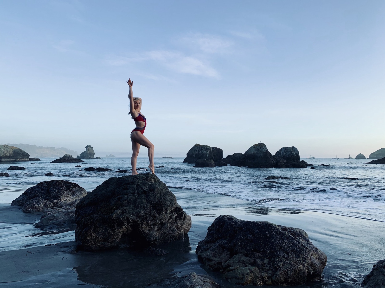 A friend in a red swimsuit poses on a large rock at a rocky beach, one arm stretched overhead.