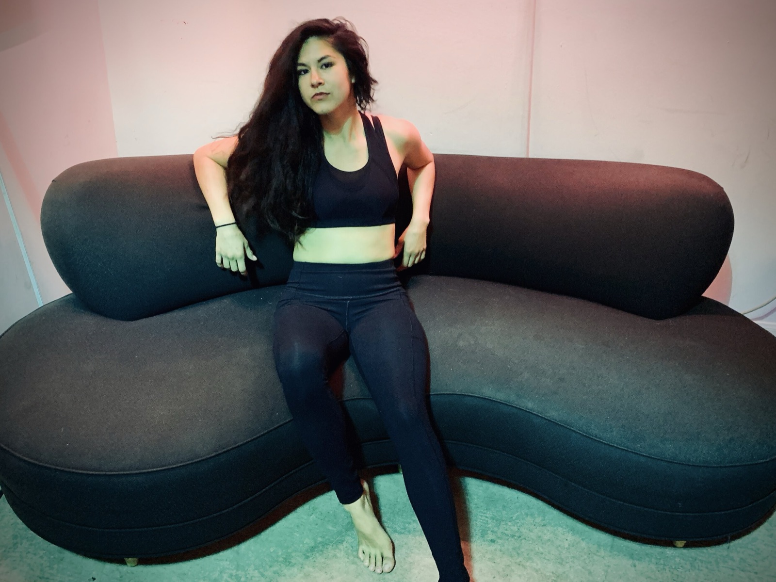 Khutulun in a black sports bra and leggings sits on a curved dark sofa against a pink-lit wall.