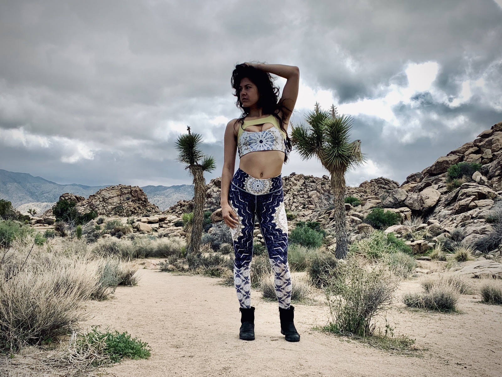 Khutulun stands on a sandy desert trail among Joshua trees and boulders, one hand raised to her hair.