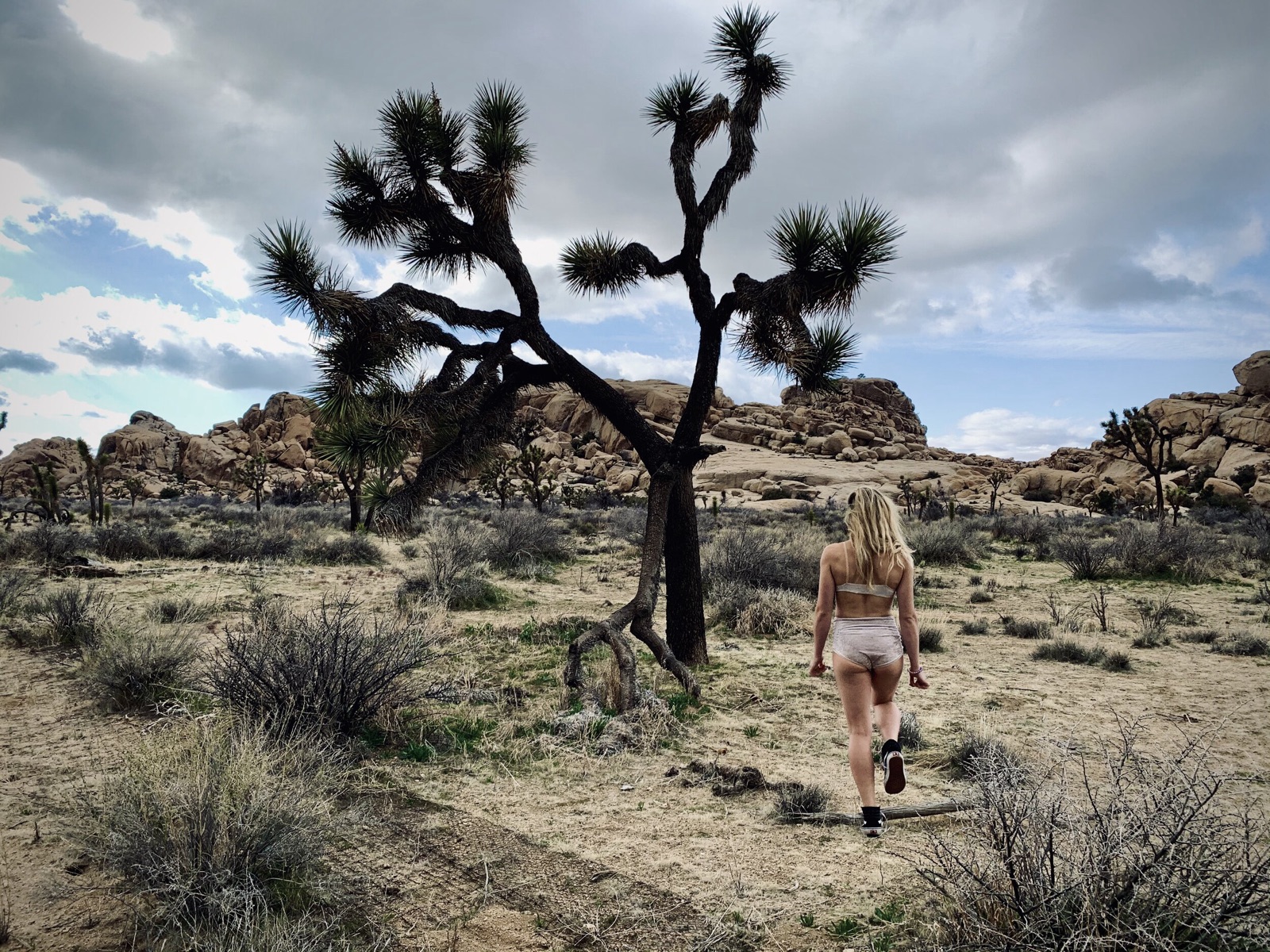A friend in a two-piece outfit walks past a Joshua tree toward desert rock formations under a cloudy sky.