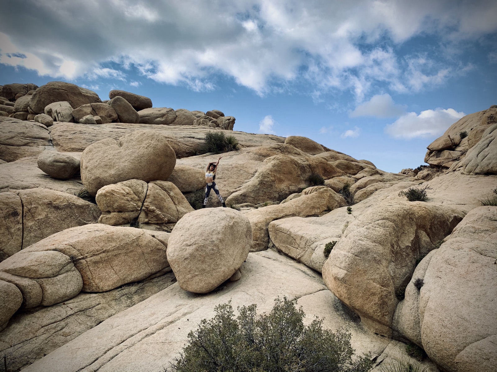 A person poses with one arm raised on a vast slope of rounded boulders under a partly cloudy blue sky.