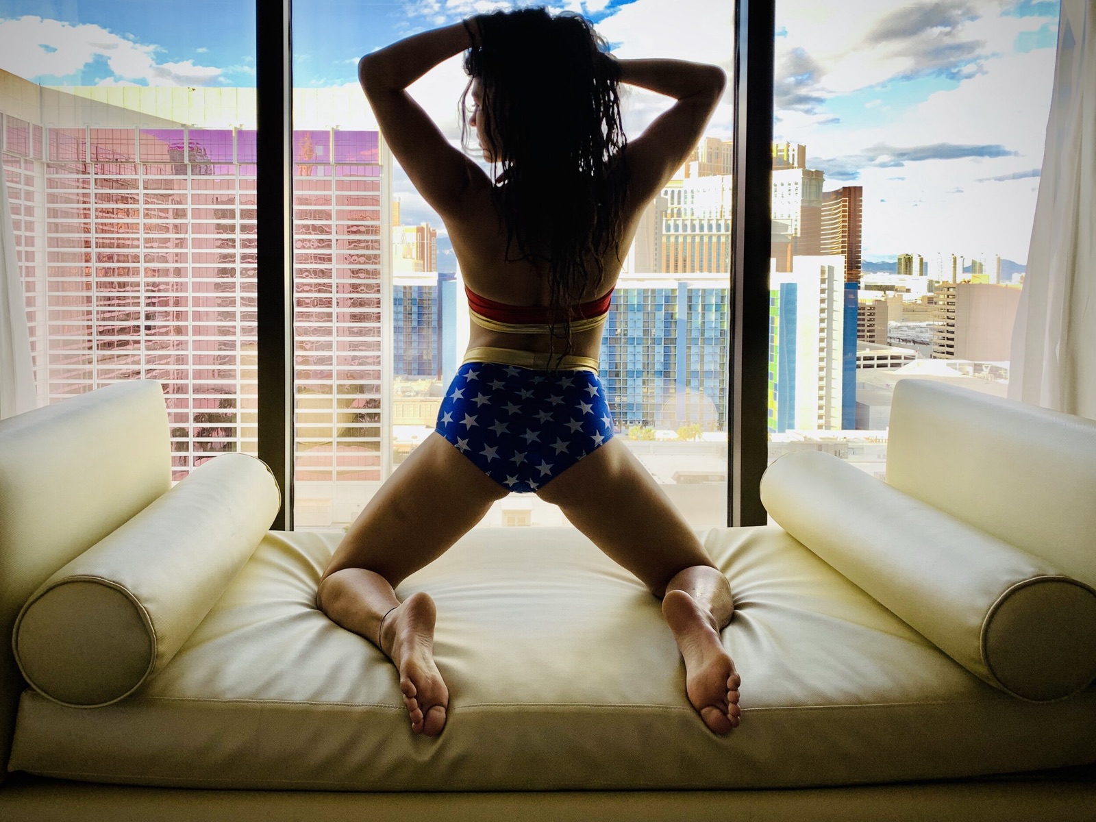 Khutulun kneels on a window bench in a Wonder Woman costume, facing a view of Las Vegas hotel towers.