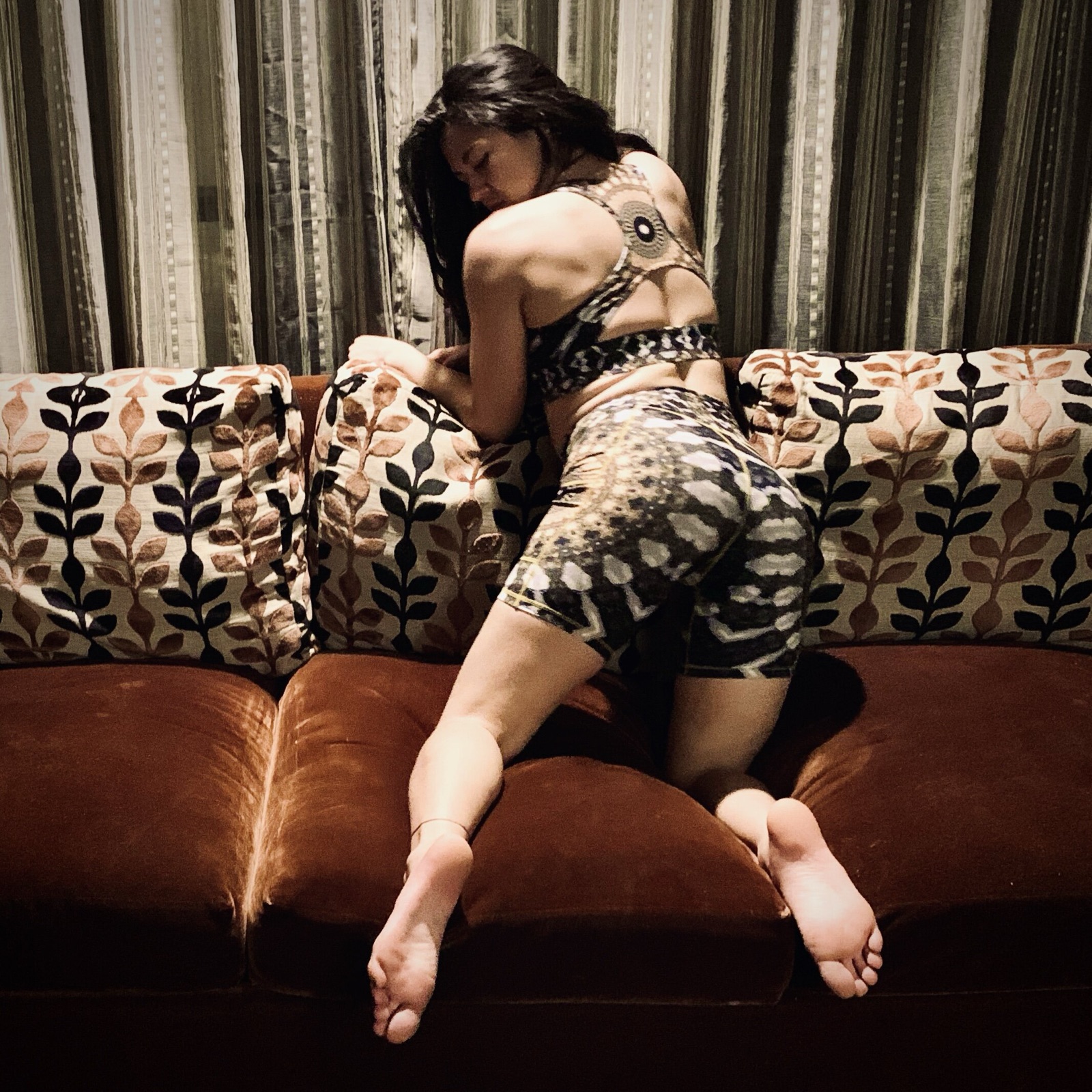 Khutulun kneels facing away on a brown velvet couch in patterned athletic wear, leaning on the cushions.