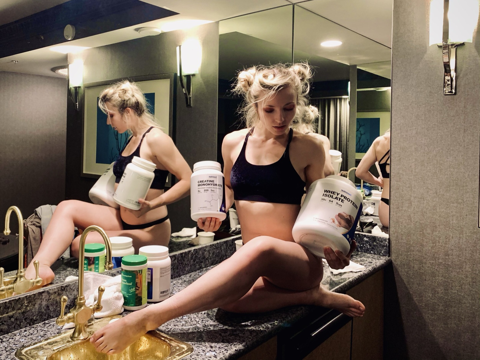 A friend sits on a hotel bathroom counter between mirrors, holding large tubs of protein and creatine powder.