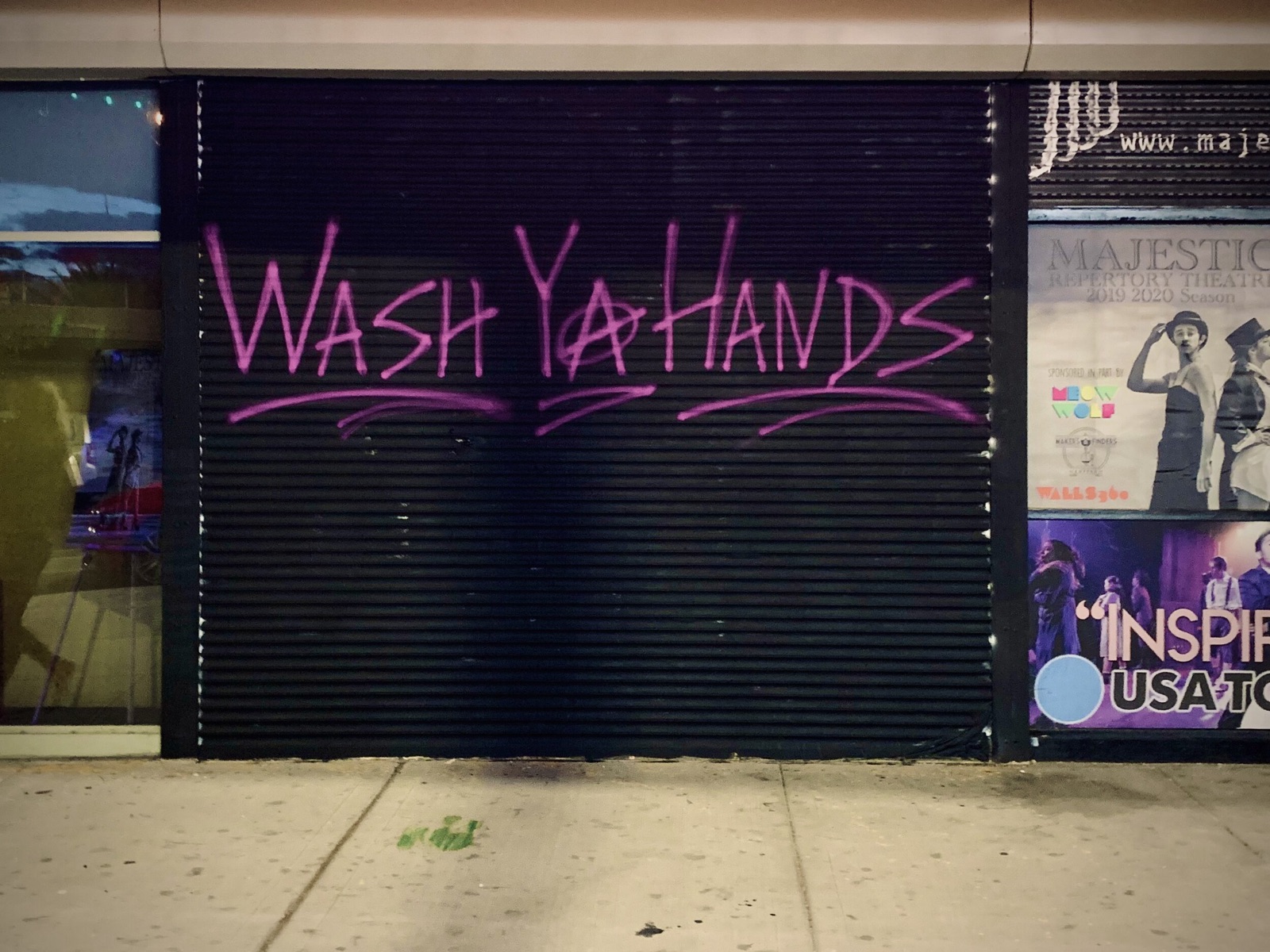 Wash Ya Hands spray-painted in pink on a dark storefront shutter beside theater posters.
