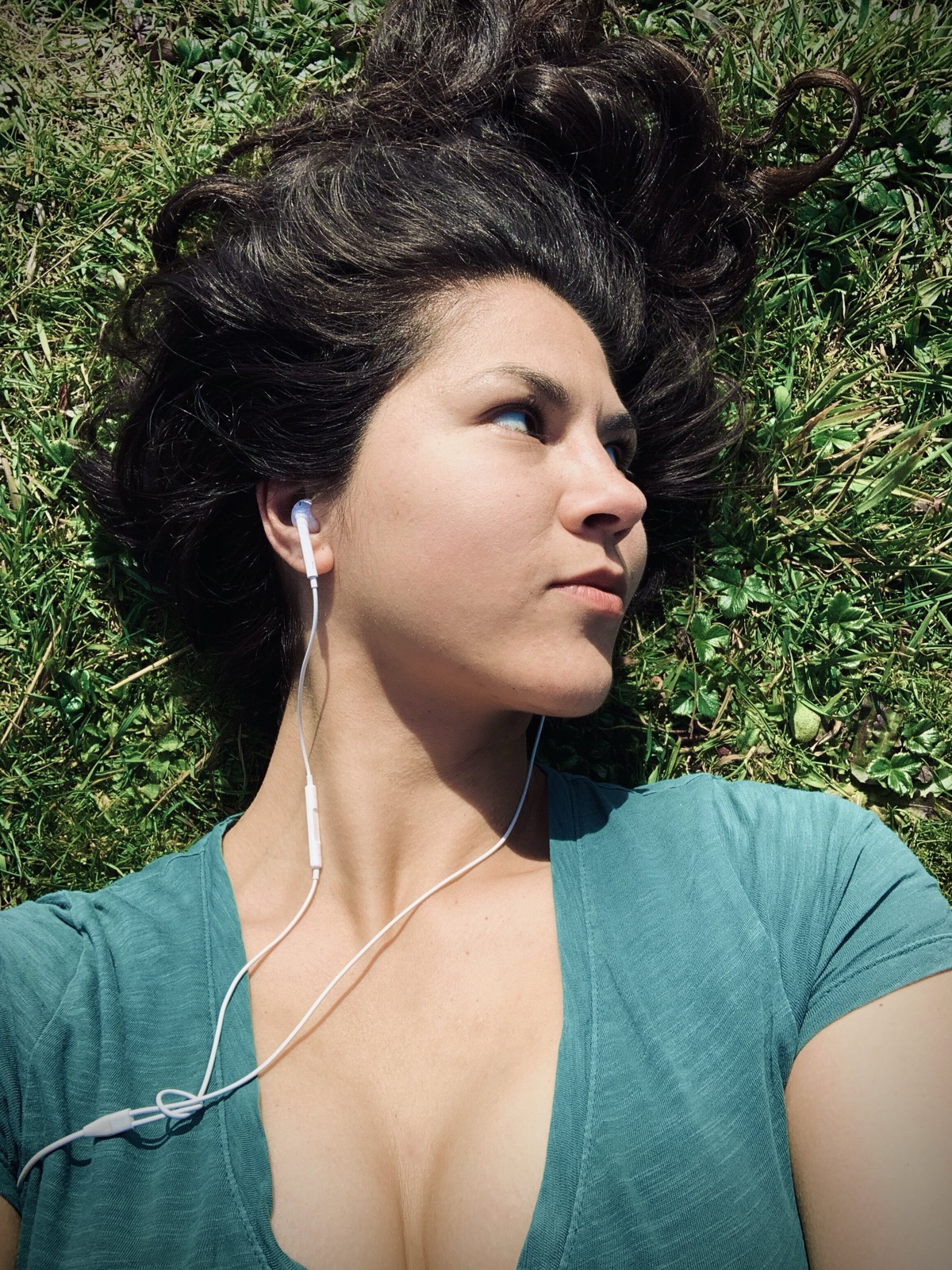 Khutulun lies in green grass wearing earbuds and a teal shirt, dark curly hair spread out, gazing to the side.