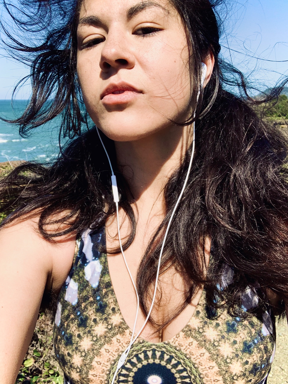 Selfie of Khutulun with windblown dark hair and earbuds, blue ocean and sky behind her.