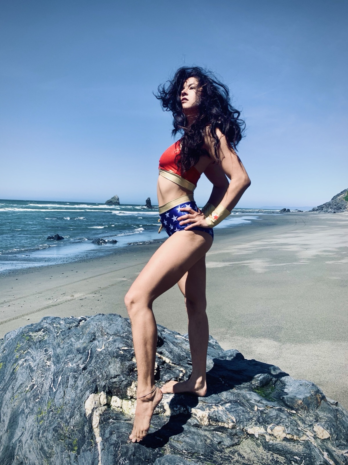 Khutulun stands on a shoreline rock in a Wonder Woman costume, wind blowing her hair.