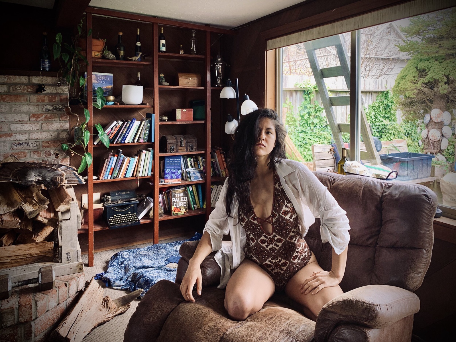 Khutulun sits on the arm of a brown recliner in a printed bodysuit and open white shirt, bookshelves behind her.