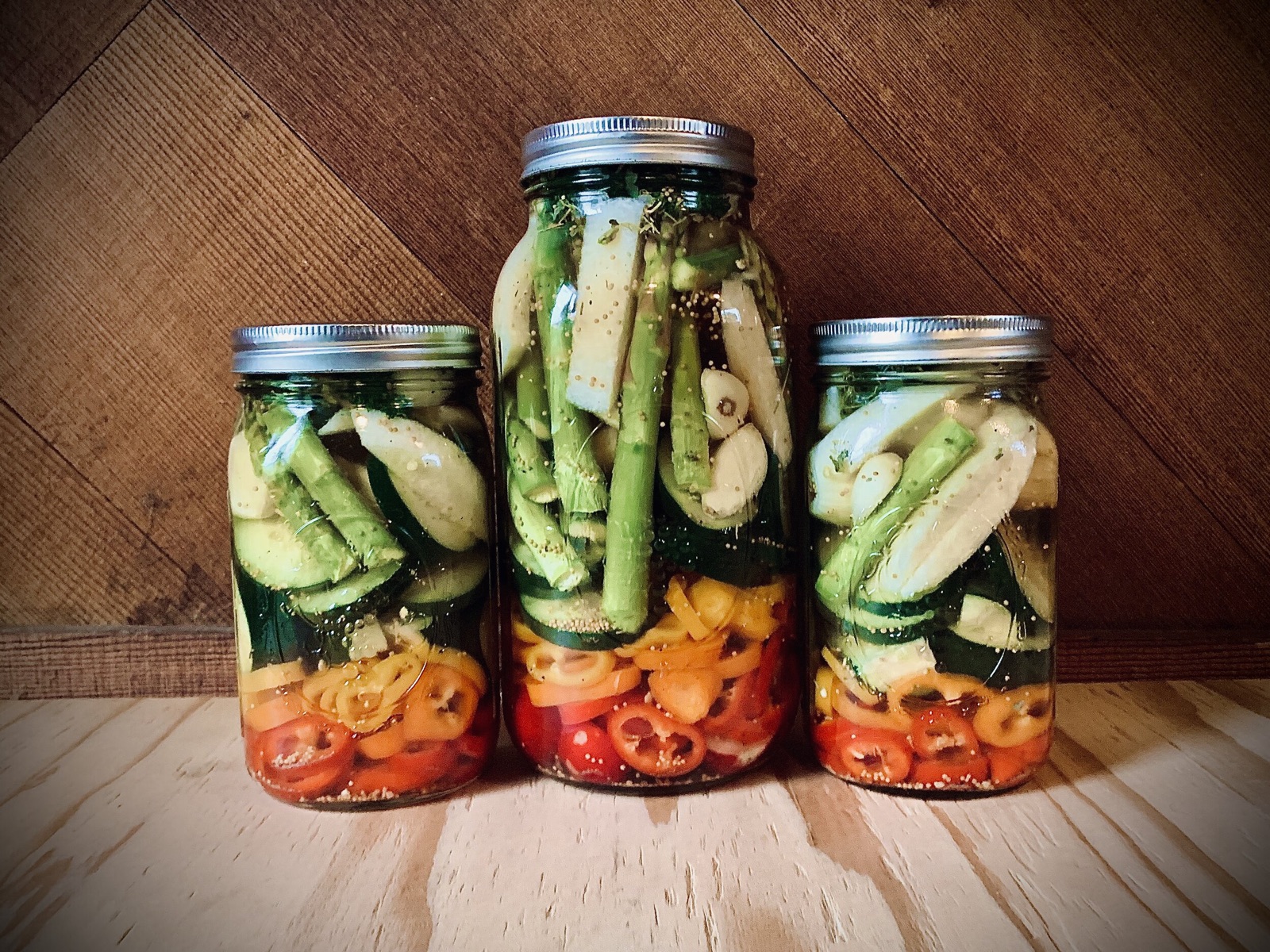 Three glass jars of pickled cucumbers, asparagus, garlic, and sliced peppers on a wooden counter.