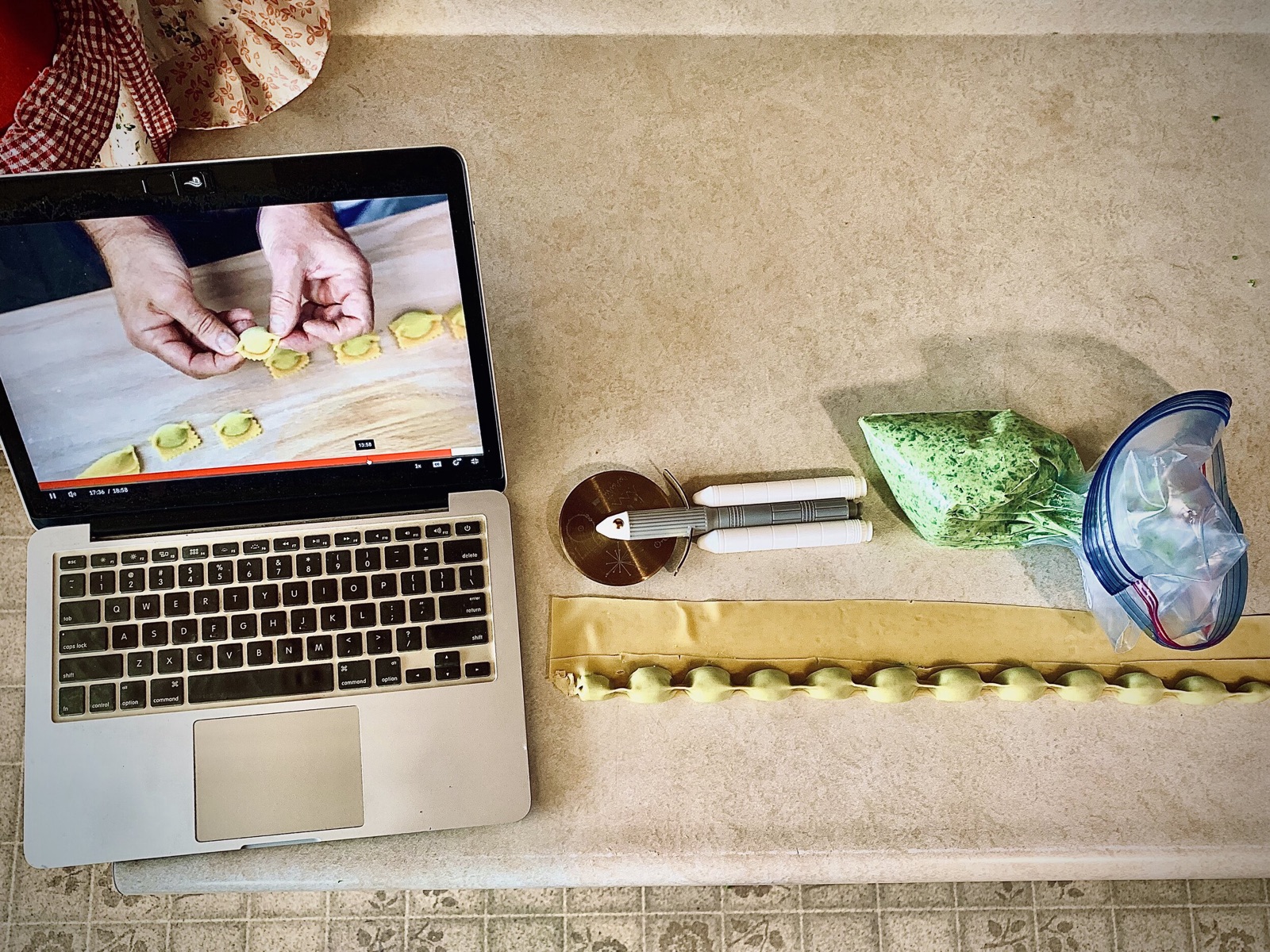 Laptop playing a ravioli tutorial on a kitchen counter beside a pastry cutter and a strip of freshly filled pasta.