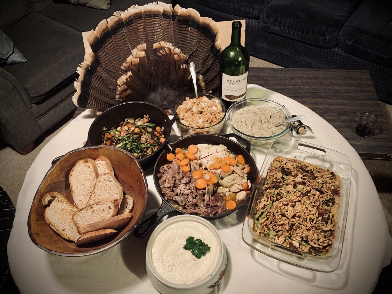 Homemade holiday dinner spread on a round table with a fanned turkey tail, wine bottle, bread, and casserole dishes.