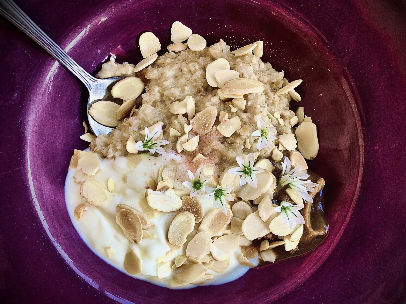 Purple bowl of oatmeal topped with sliced almonds, yogurt, nut butter, and small white flowers.