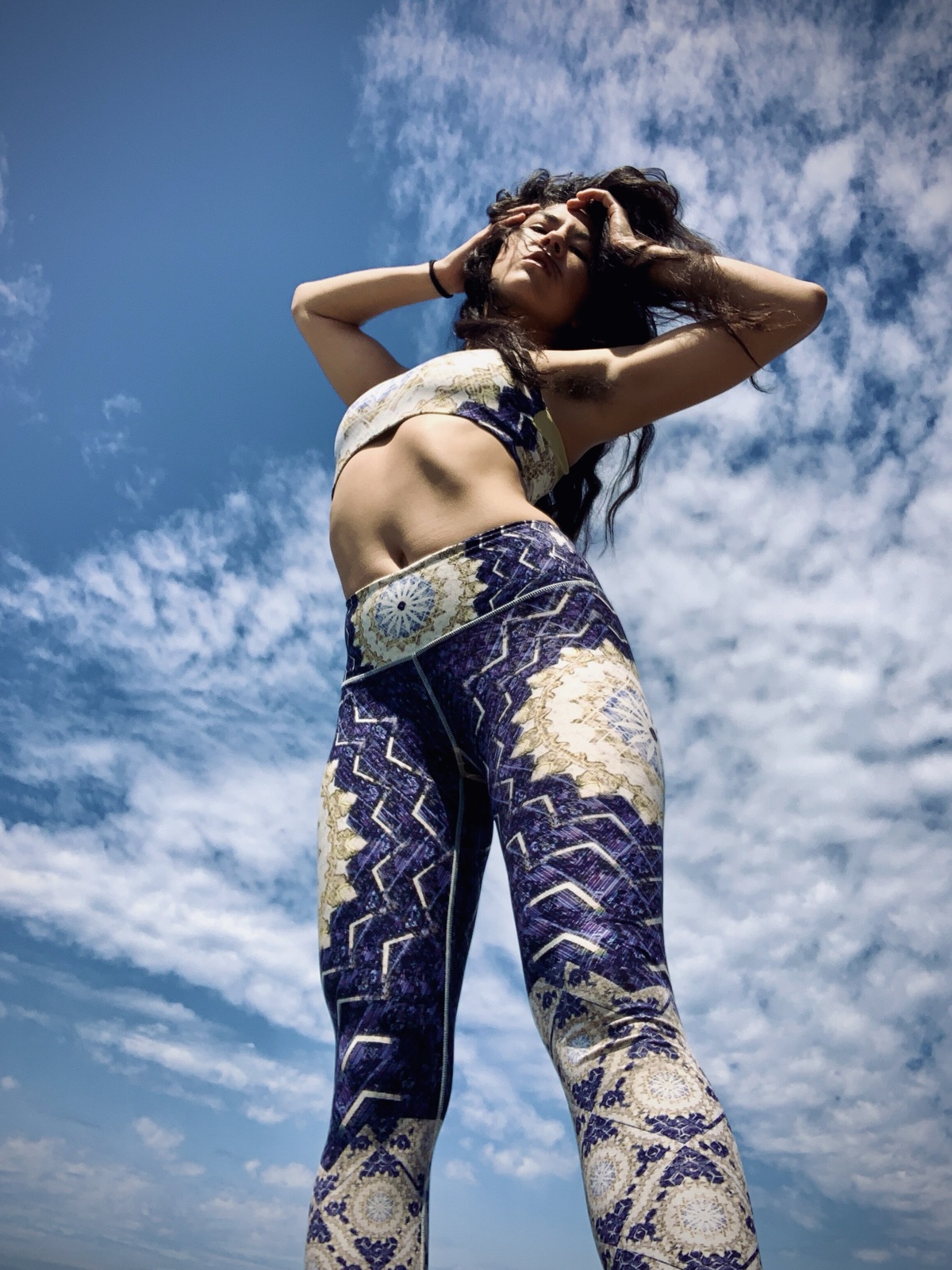 Low-angle shot of Khutulun in patterned crop top and leggings, hands in her hair against a cloud-streaked blue sky.