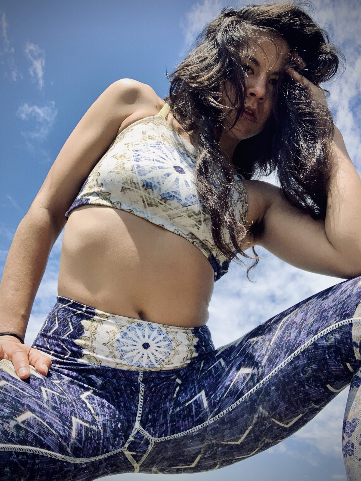 Low-angle view of Khutulun crouching in a mandala-print top and leggings, one hand in her wind-tossed hair, sky behind.