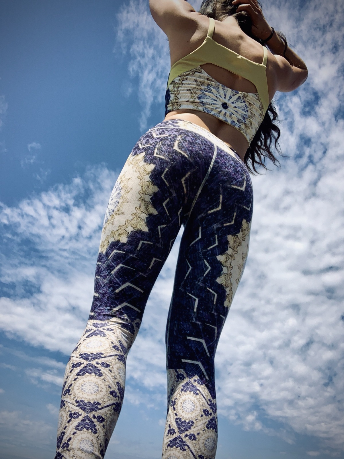 Khutulun seen from behind and below in patterned leggings and a yellow-backed top, standing against a cloudy blue sky.