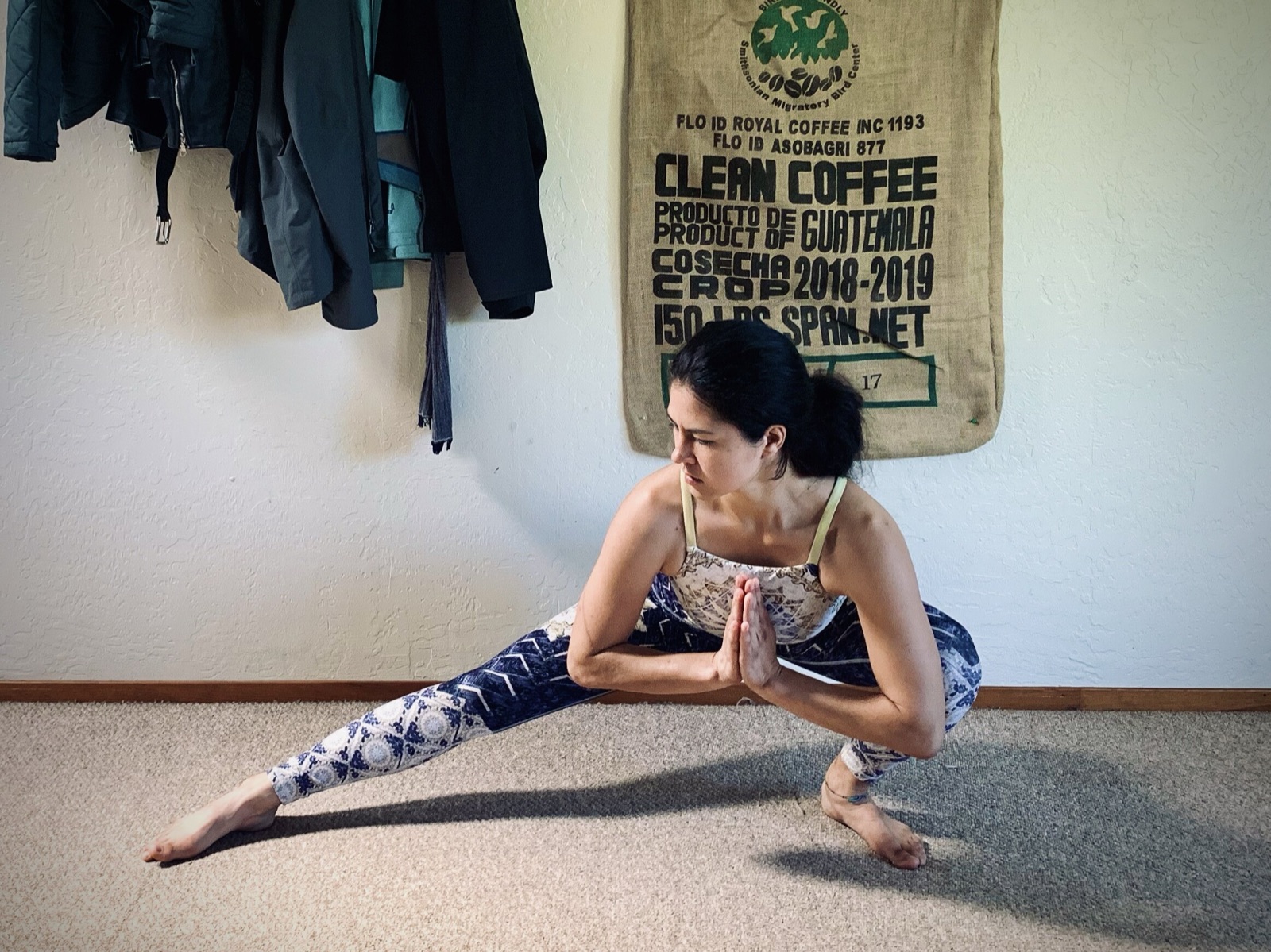 Khutulun holds a deep side-lunge stretch with palms pressed together indoors, a burlap coffee sack hanging on the wall.