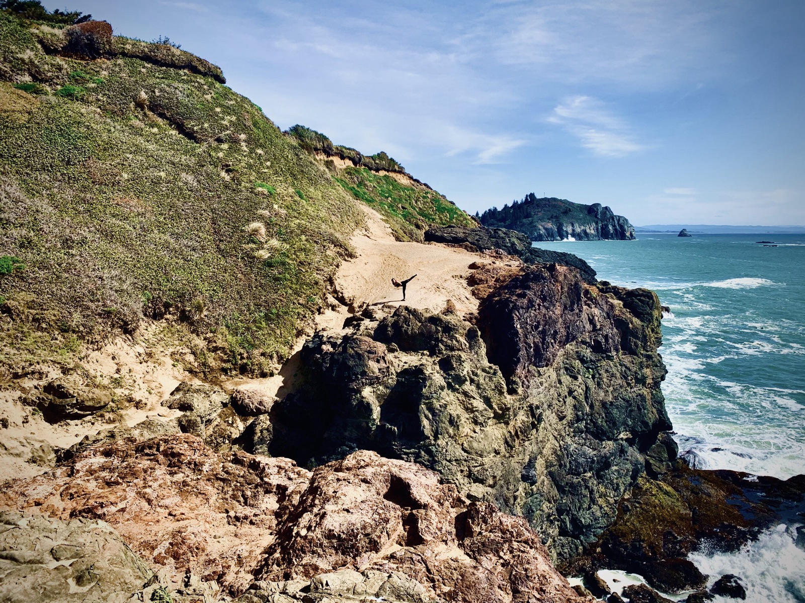 Rocky coastal cliffs and blue surf, with a distant figure balancing in a high side kick on a sandy ledge.