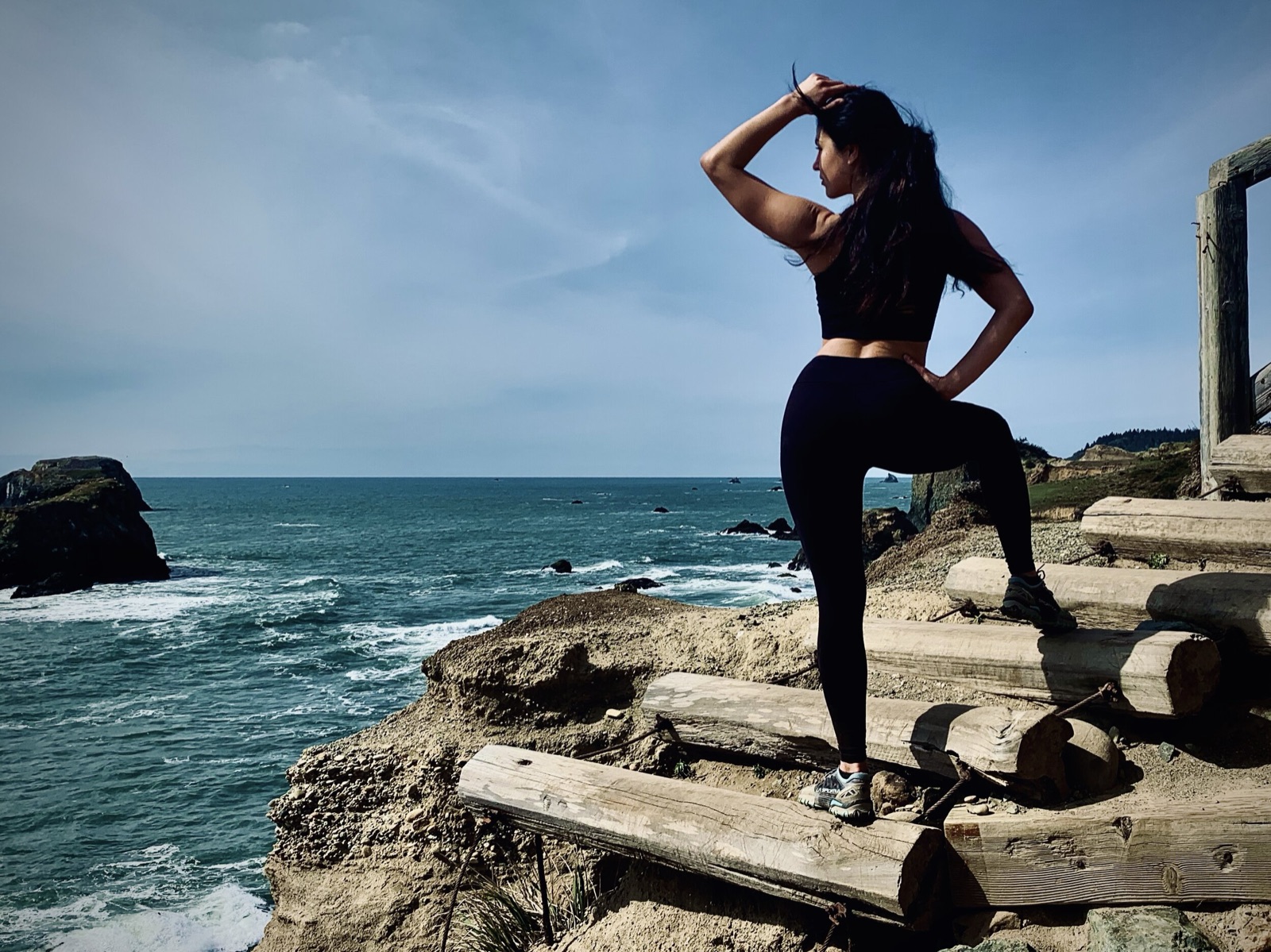 Khutulun stands on log steps above a rugged coastline in black workout wear, back to camera, hand raised to her hair.