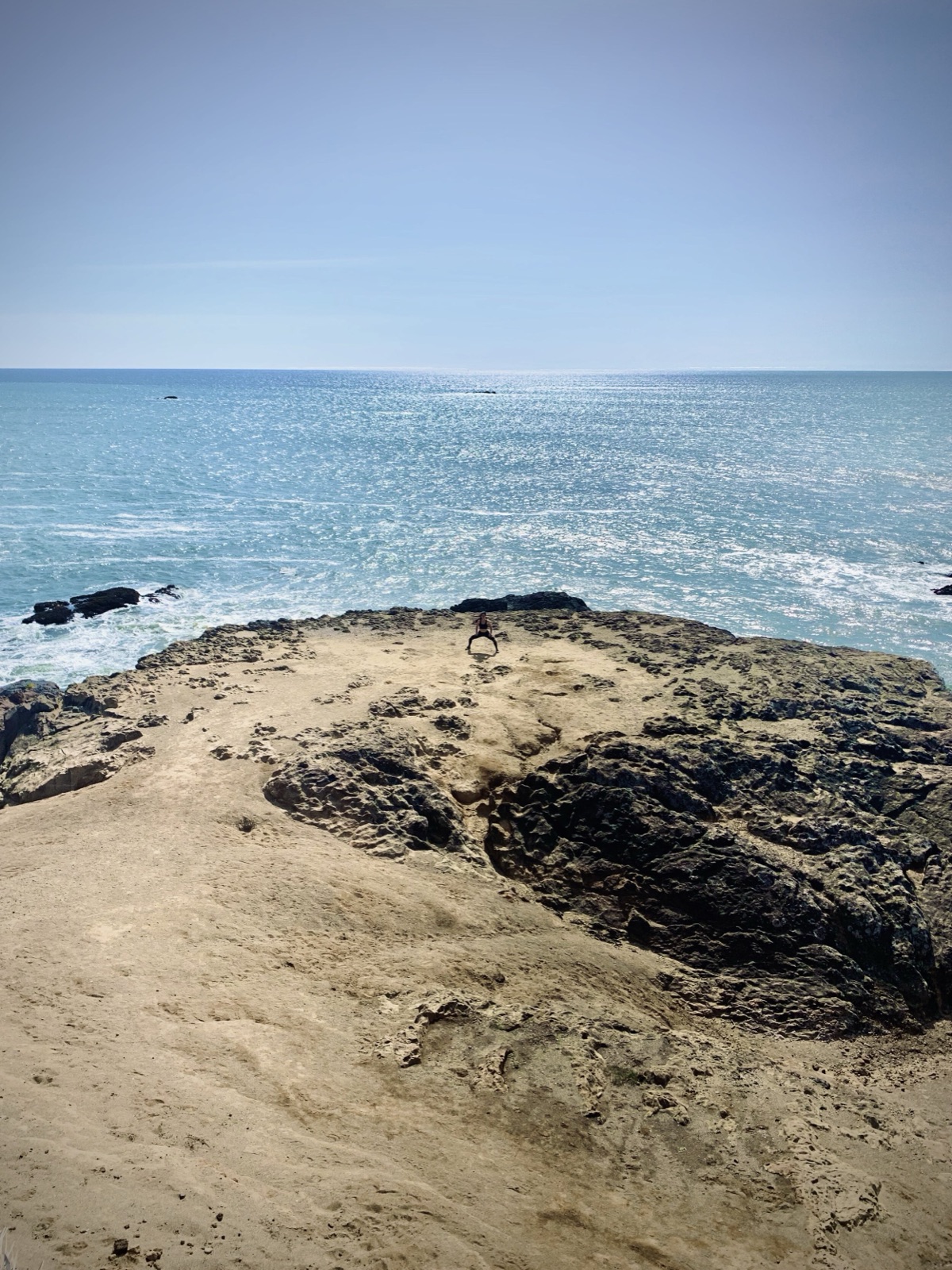 Wide view of a sandy rock outcrop over the ocean, with a tiny figure in a low squat near the edge.