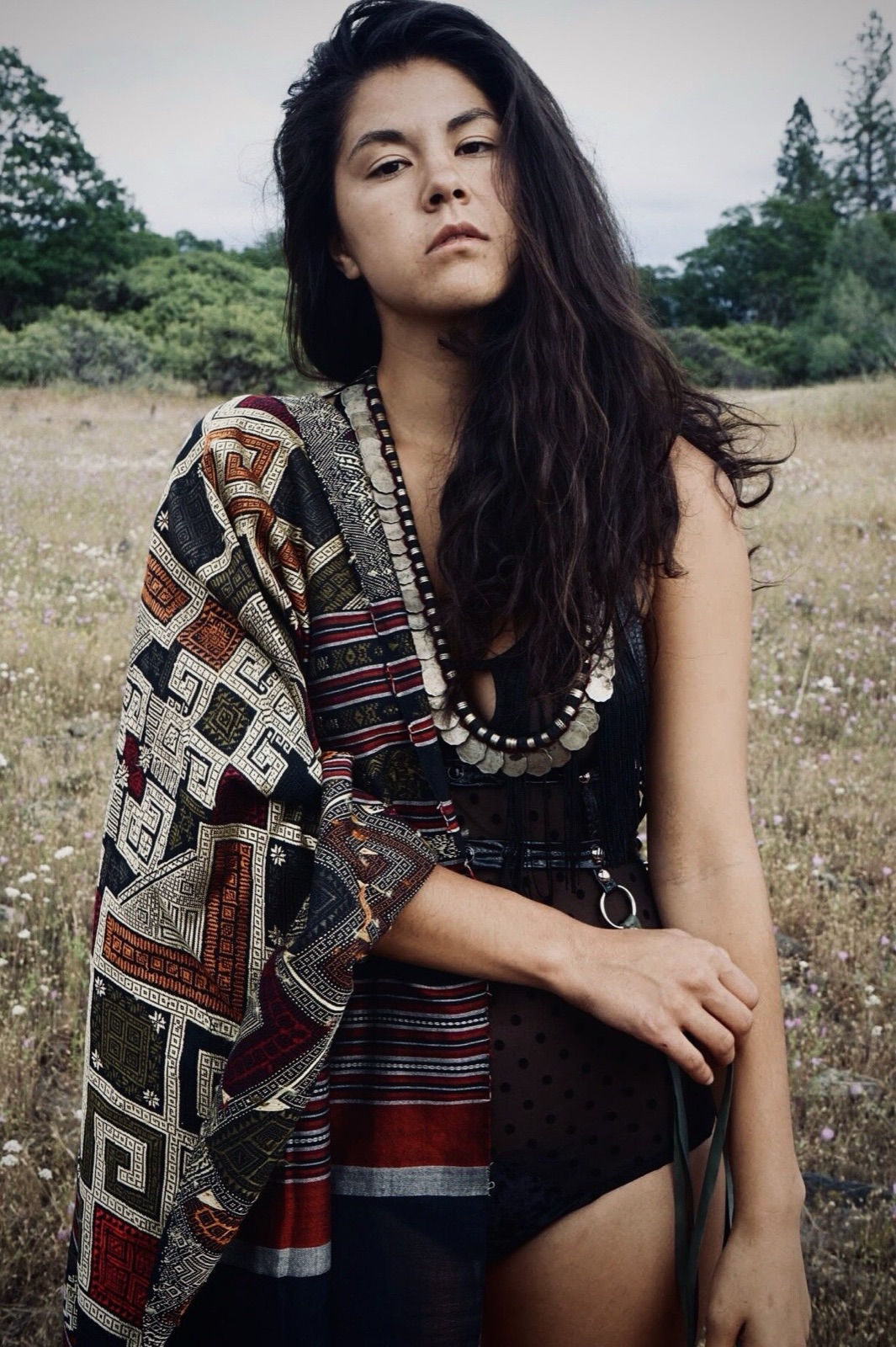 Khutulun in a wildflower field wearing a geometric-patterned shawl, layered coin necklaces, and a sheer black bodysuit.