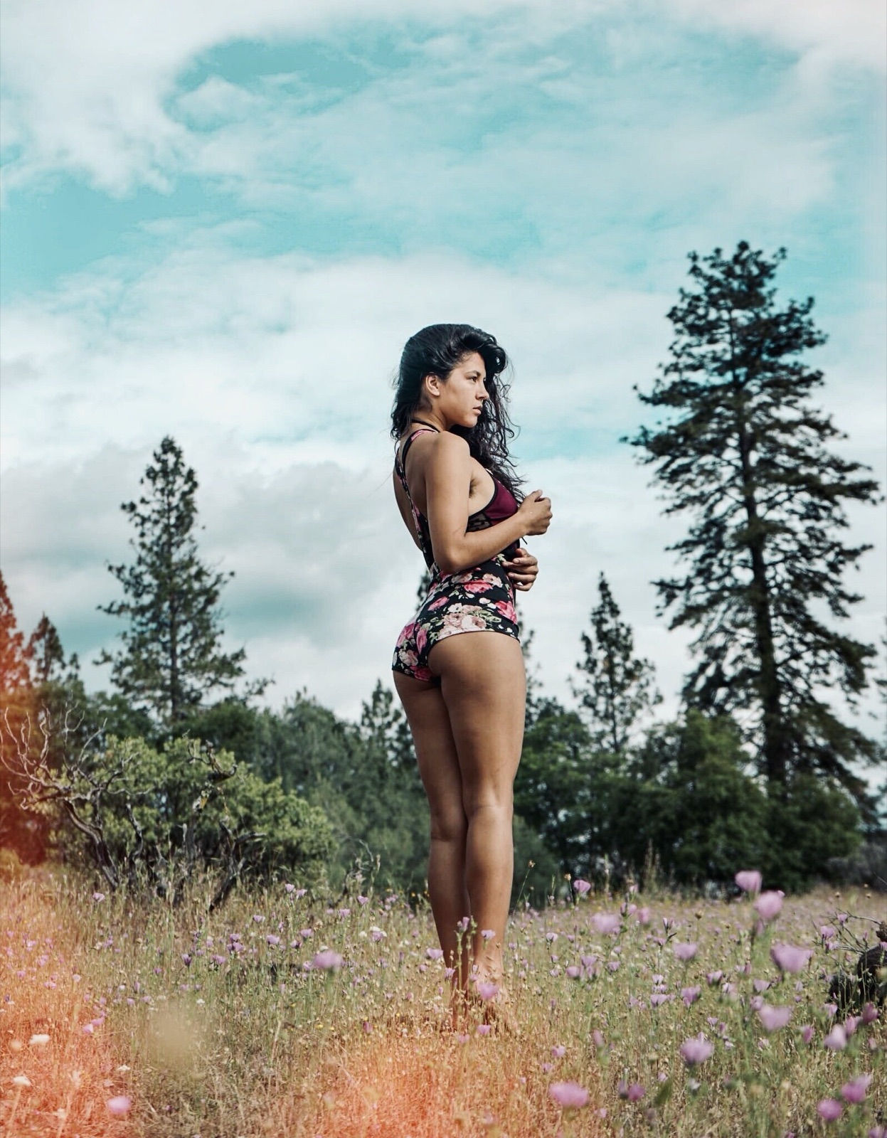 Khutulun stands barefoot in a wildflower meadow ringed by pines, wearing a floral one-piece and looking into the distance.